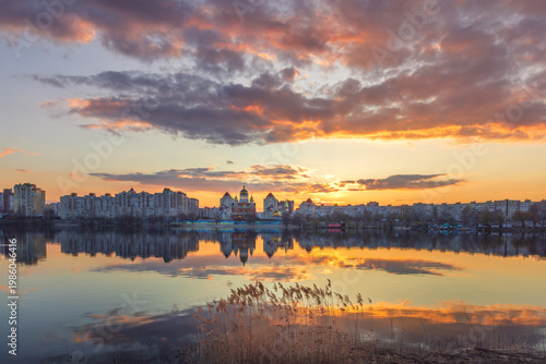 Golden sunset melts over Obolon embankment in Kyiv, painting the Dnipro River with shimmering reflections. Clouds drift softly as glowing domes and skyline dissolve into a calm, dreamy evening.