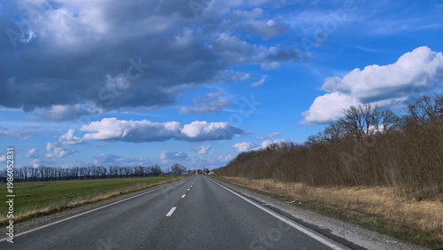 View from a car window of a straight road with white lane markings under a blue sky with clouds. Concept of travel, journey, movement forward, and freedom. Transportation, road trip, and perspective.