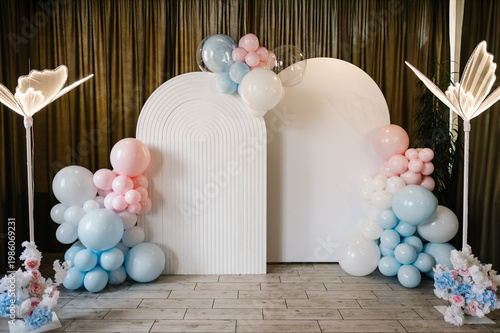 Elegant photo zone for a baby shower or birthday party featuring two white arch backdrops and blue, pink, and white balloon. Modern event decoration with floral arrangements and luminous butterflies.