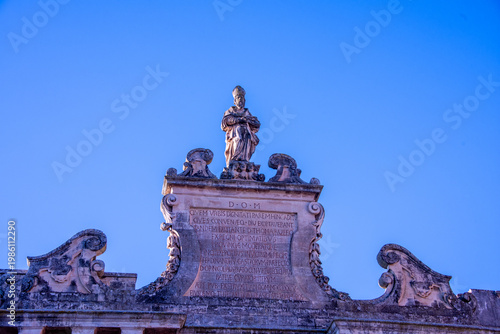 The upper section of Porta San Biagio in Lecce, featuring a statue of Saint Blaise above an inscribed pediment, framed by elegant baroque scrolls against the clear sky.
