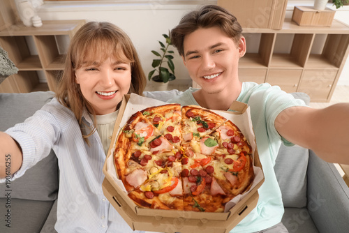 Beautiful young couple holding cardboard box with tasty pizza and taking self...