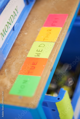 Organised production trolley with labelled plastic bins for each weekday in a bright factory setting, vivid blue structure with colourful labels creating a structured orderly atmosphere