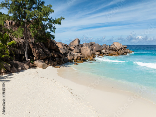 Granite rocks meet the turquoise ocean at a white sand beach, surrounded by lush greenery. Grand Anse Beach. Seychelles, La Digue.