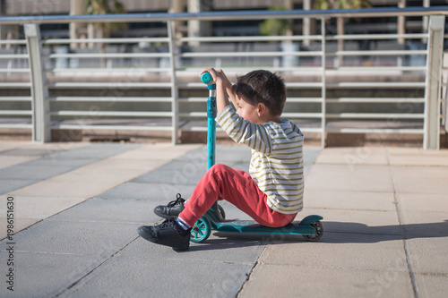 A cheerful five-year-old boy sits on a turquoise scooter, playing on a smooth, paved surface in a vibrant urban environment