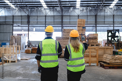 Two Caucasian workers conduct detailed warehouse inspection, pointing at machinery amidst stacked wooden pallets and boxes in a bright, industrial factory setting, wearing high-visibility yellow vests
