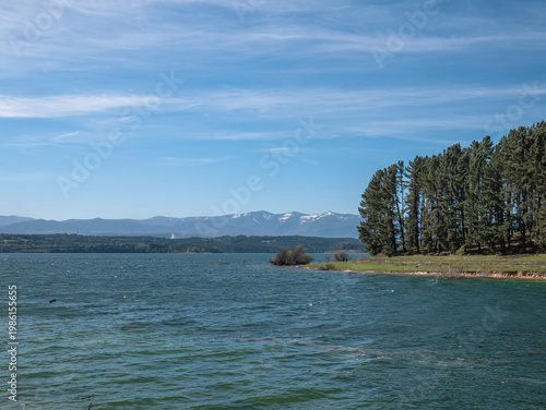 View of the shore of the Barcena Reservoir full of very tall pine trees close to the small waves caused by the wind and some mountains of Bierzo with snow on the peaks at the beginning of spring