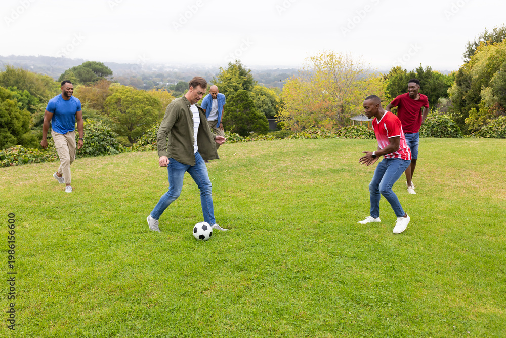 Obraz premium Diverse male friends playing soccer on lawn, dribbling black-and-white ball, wearing green jacket