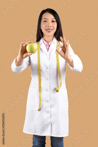 Female Asian nutritionist with apple showing OK on beige background