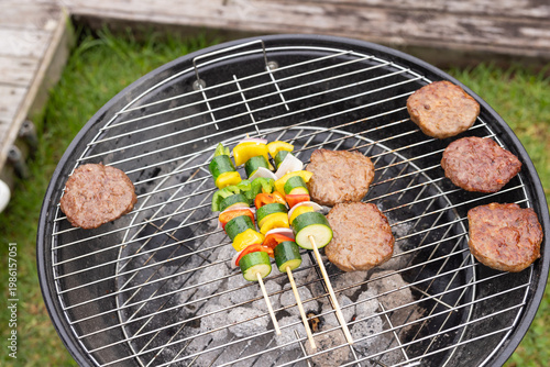 Charcoal kettle grill sitting on lawn near deck, holding six beef patties and three veg skewers
