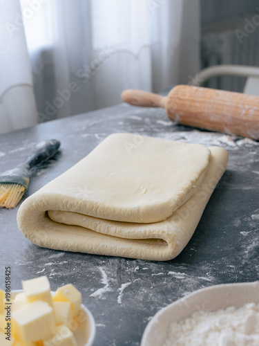 A fold raw puff pastry dough, with butter, flour and rolling pin on table. It's a laminate process to creates multiple alternating layers. Pastry composed with dough and butter folding.