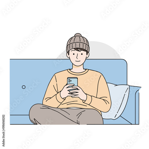 A young man wearing a beanie sits cross-legged on a blue couch, absorbed in his smartphone, with a pillow nearby.