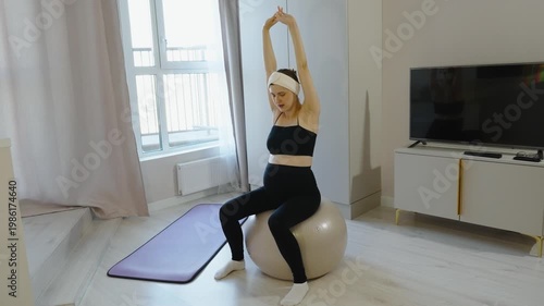 Expecting mother, takes care of her body, doing morning exercises on fitball in her apartment. Pregnant woman sits on ball, stretches muscles of her arms and body, and tones body before giving birth.
