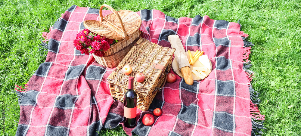 Fototapeta premium Wicker baskets with flowers, wine and food for picnic on plaid in park