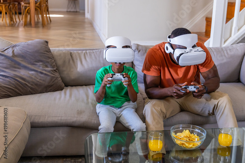 African American father and son playing VR on sofa wearing headsets with controllers, chips, juice