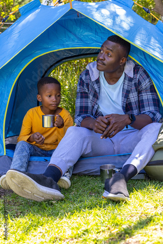 African American father and son sitting at tent entrance holding metal mugs, backpack nearby