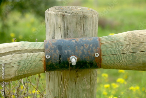 Detalle de valla de madera rústica con unión metálica, cuya forma sugiere un ave imaginaria con alas abiertas en un entorno natural
