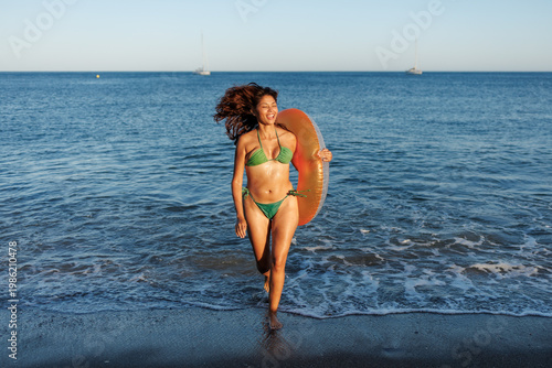 Obraz na płótnie Woman in green bikini running from the ocean with an orange inflatable ring