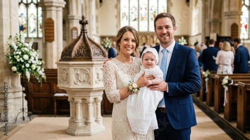 A smiling couple holds their newborn baby during a baptism ceremony in a historic church. The woman wears a lace wedding dress and a blue tie, while the man wears a navy suit.