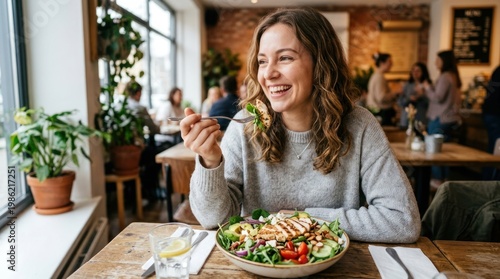 A smiling woman enjoys a salad at a restaurant table. She is seated at a wooden table with a leafy salad in front of her.