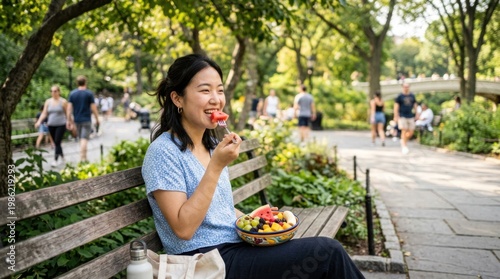 A young woman enjoys a refreshing summer day in a park. She sits on a wooden bench, eating a slice of watermelon with a spoon from a colorful fruit bowl.