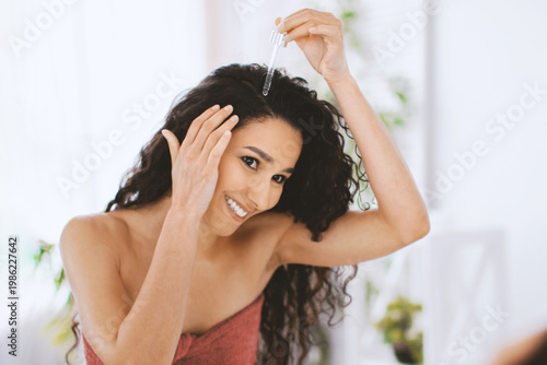Tableau sur toile A woman is using a dropper to apply hair oil to her scalp while looking in the mirror
