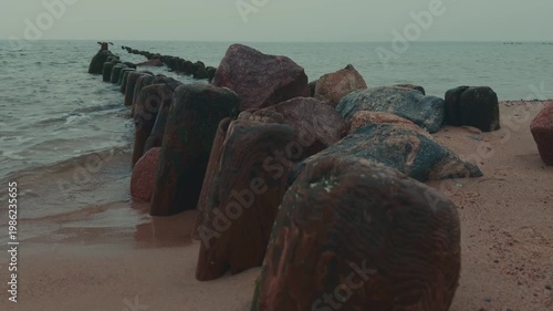 Wooden groynes and large rocks on the seashore. Coastal landscape with rough waves hitting barrier structures. Minimalist marine scenery for nature background.