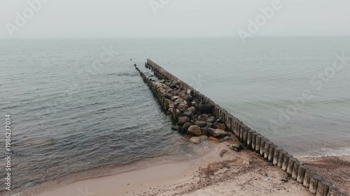 Wooden groynes and large rocks on the seashore. Coastal landscape with rough waves hitting barrier structures. Minimalist marine scenery for nature background.
