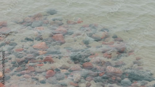 Clear river water flowing over multicolored pebbles on a sandy bottom. Close up view of stones submerged under rippling current. Natural aquatic background scene.