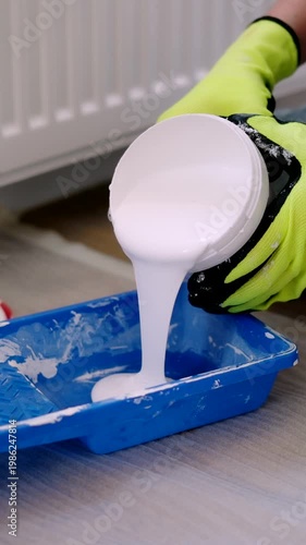 House Painter in gloves pours white paint from Can into a blue tray At Living Room Renovation Painting