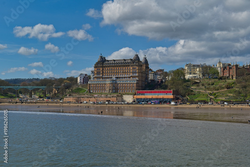 A grand hotel overlooks a sandy beach and the sea under a partly cloudy sky. People enjoy the beach on a sunny day in Scarborough - Yorkshire - Great Britain
