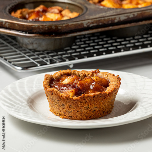 A mini apple pie tart on a plate with a pan of the rest cooling in behind.