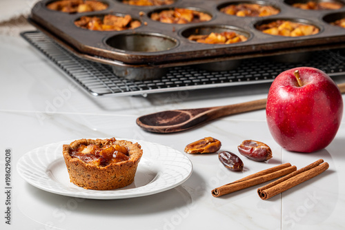 A mini apple pie tart with the rest in a muffin tin in behind and ingredients.