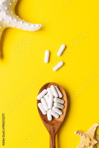 Wooden spoon with white medical capsules of sea collagen on bright yellow background decorated with decorative starfish. Health care products.