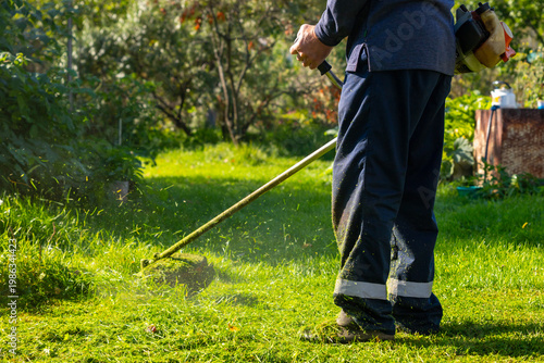 Man using grass trimmer to cut lawn in backyard. Gardener operating string trimmer to maintain greenery. Lawn care equipment, landscaping tool and grass maintenance gardening concept.