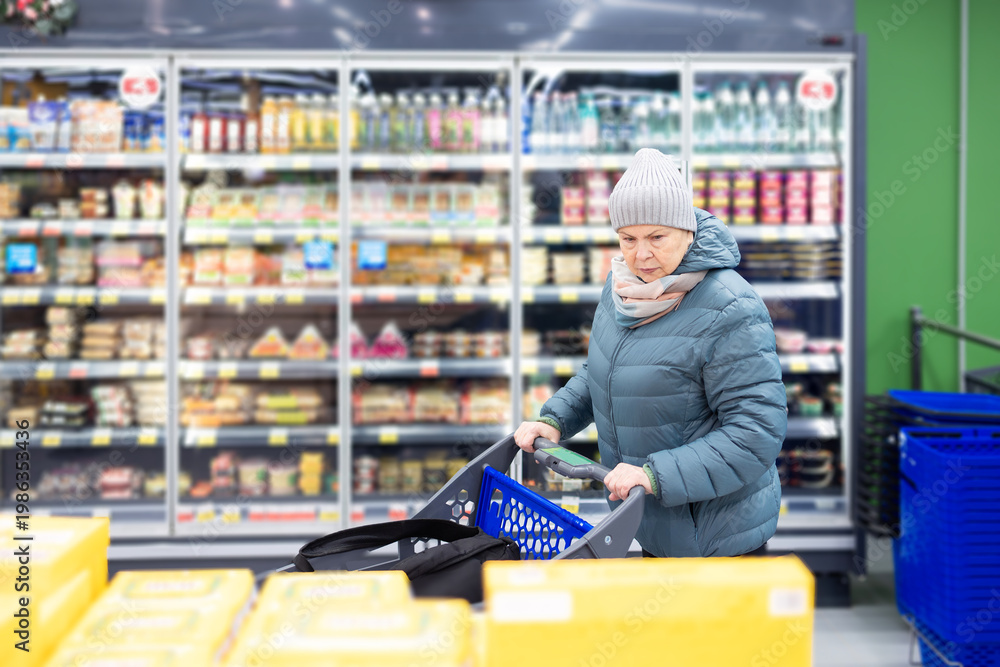 Obraz premium Mature woman in warm jacket and knit hat pushing shopping cart inside brightly lit supermarket aisle with frozen food section.