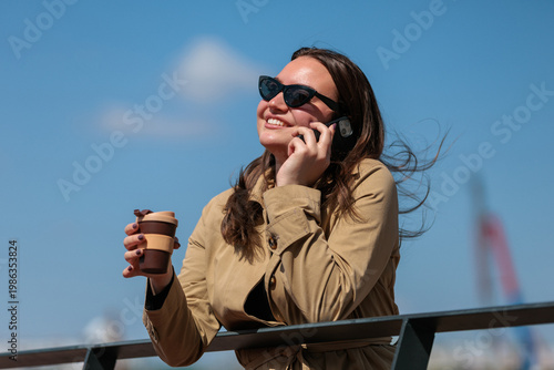 Young woman talks on phone while enjoying coffee on a sunny day at an outdoor location near water