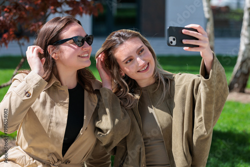 Two young women take a selfie together while sitting on a sunny day outside in a green park area
