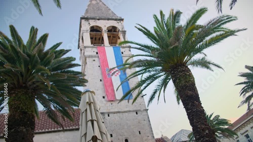 morning establishing shot of the 14th-century bell tower of the Church and Monastery of St. Dominic in Trogir, Croatia. The stone tower features a Croatian flag, framed by lush palm trees