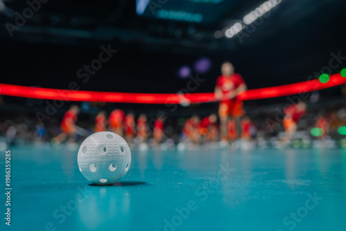 Close-up of a white floorball on a turquoise court with players in red uniforms blurred in the background
