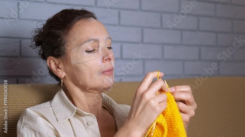 Woman knitting and relaxing with facial sheet mask. Relaxed woman sitting on a comfortable sofa knitting with bright yellow yarn while wearing a cosmetic face mask for skincare, enjoying her hobby