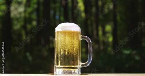 Freshly brewed craft lager bear with foam. Beer mug in summer nature. Sparkling amber ale beer with natural background. Barley malt craft beer on wooden table. Pouring fresh beer with splash.