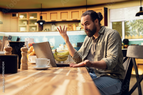 Фотография Uncertain freelancer being confused about ambiguous tasks at home, remote work on laptop in the kitchen