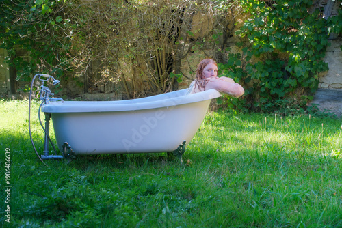 Redhead woman bathes in vintage tub placed outdoors on grass surrounded by greenery while looking back creating unusual scene combining nature and hygiene with quiet reflective mood