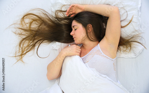 Young woman sleeps on white bed with long hair spread across pillow wearing light clothing while gentle shadows and soft setting create quiet restful intimate atmosphere