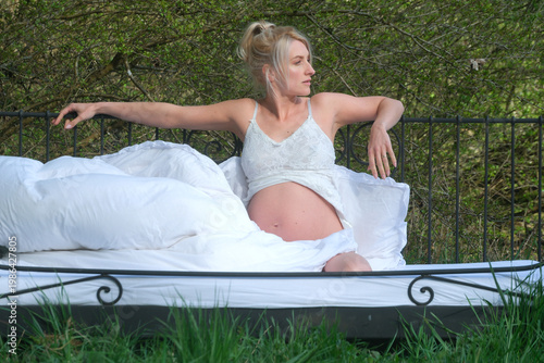 Pregnant woman sits on bed outdoors surrounded by greenery wearing light clothing while resting and looking aside creating natural intimate scene with calm maternal atmosphere