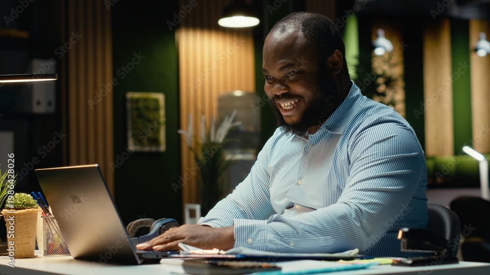 Fototapeta premium Excited african american man receiving good news for career opportunity, being proud of his breakthrough and accomplishment in the office late at night. Delighted about new deal.