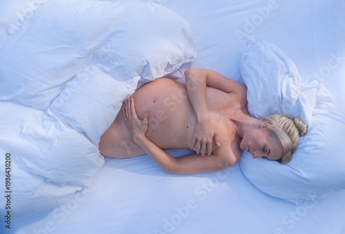 Pregnant woman sleeps on white bed gently holding belly with soft blue tones and light bedding creating intimate peaceful scene focused on rest and maternal connection
