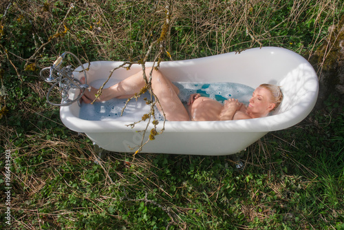 Pregnant woman relaxes in vintage bathtub outdoors surrounded by grass and branches holding belly while resting in water creating unusual natural setting with intimate serene mood