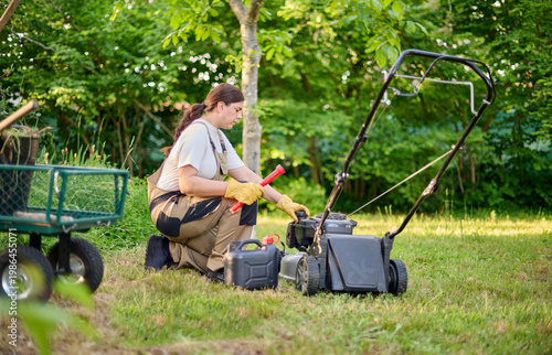 Professional female gardener prepares to refill the gasoline tank of a lawn mower beside a utility cart, surrounded by gardening machinery and tools in an outdoor work environment