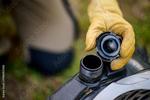 Close view of a hand with work glove holding the fuel cap of a lawn mower, preparing to add gasoline before starting gardening tasks in an outdoor maintenance setting